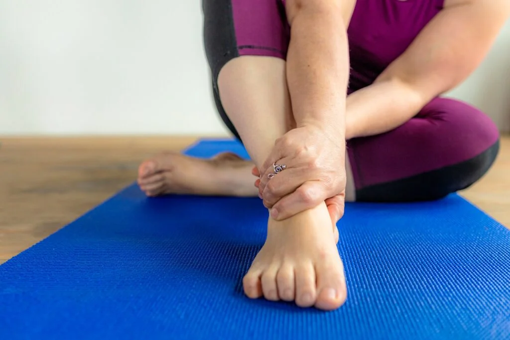 Person holding their ankle while sitting on an exercise mat, representing acute foot and ankle pain or injury.